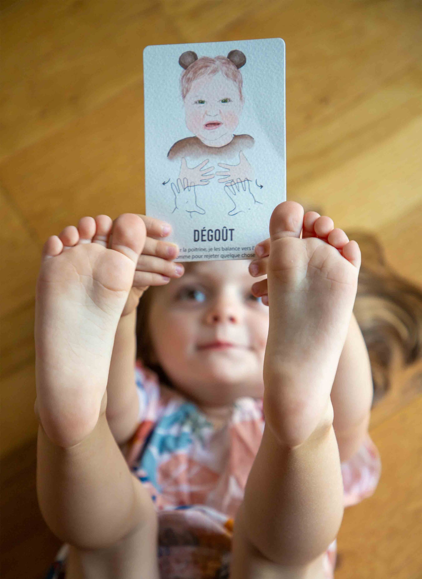 Enfant tenant la carte de signe bébé "dégoût", allongée sur le parquet, les pieds et les mains tendus vers le ciel.